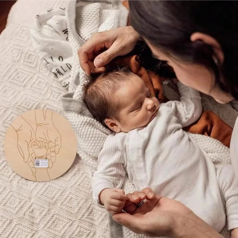Newborn baby being held by a person with a wooden disc featuring handprints in the corner.