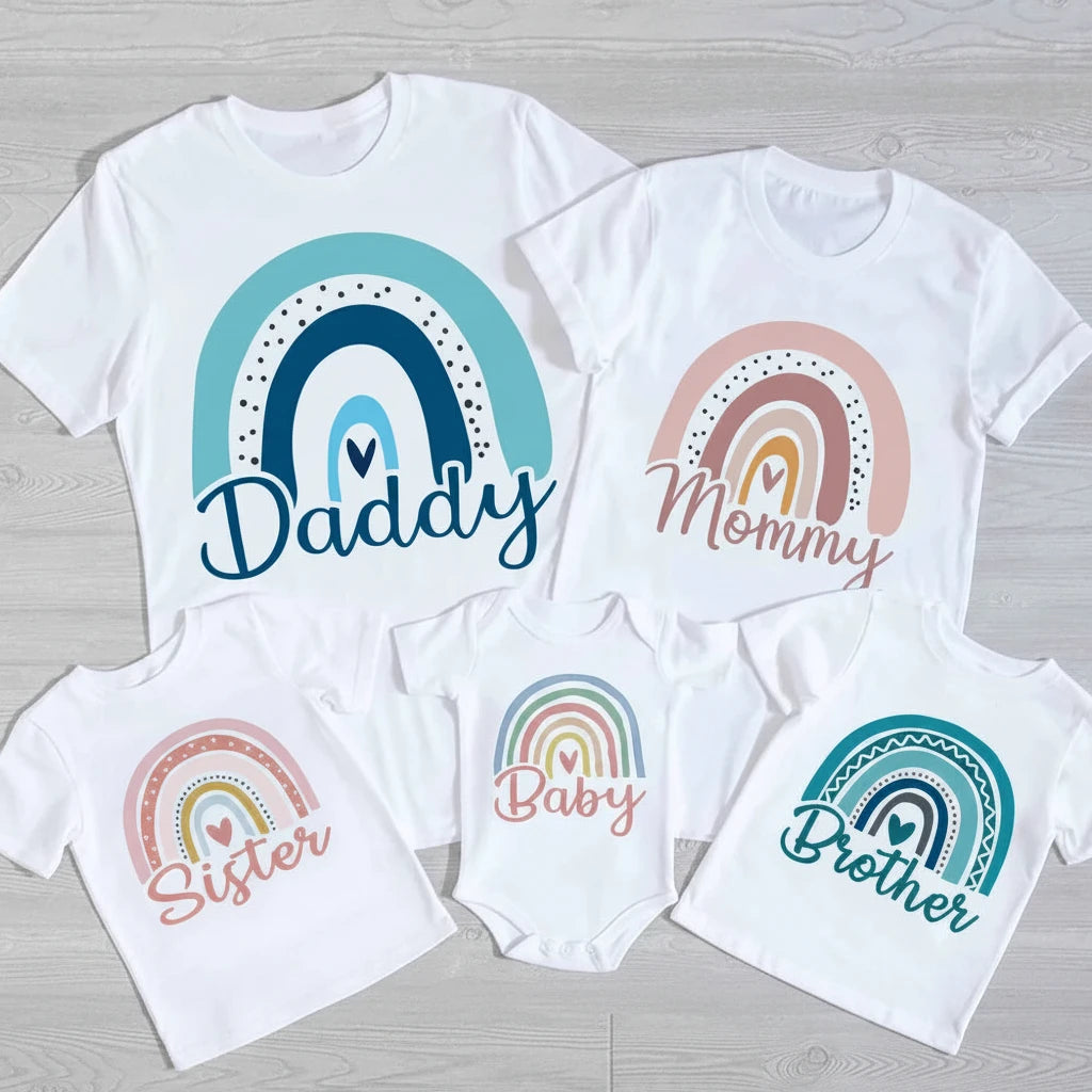Set of white t-shirts with rainbow designs and text for 'Daddy', 'Mommy', 'Baby', and 'Brother' on a wooden surface.
