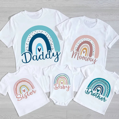 Set of white t-shirts with rainbow designs and text for 'Daddy', 'Mommy', 'Baby', and 'Brother' on a wooden surface.