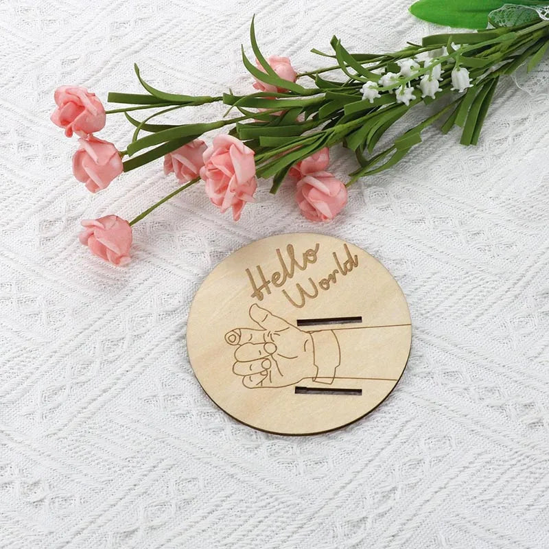 Wooden disc with 'Hello World' and handprints on a white background with pink flowers and green leaves.