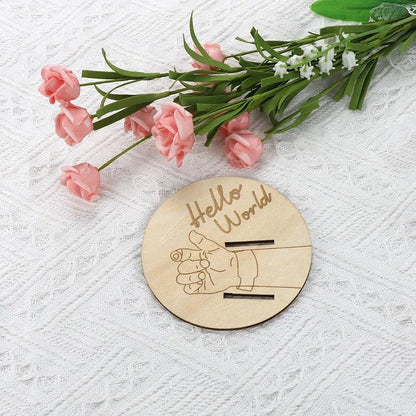Wooden disc with 'Hello World' and handprints on a white background with pink flowers and green leaves.