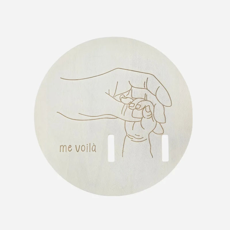 Round beige product with a line drawing of a hand holding a baby and 'me voila' text on a light gray background