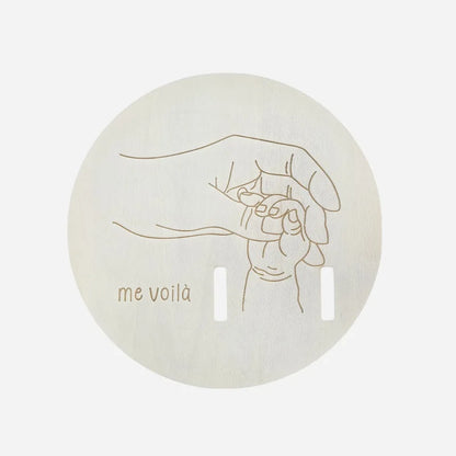 Round beige product with a line drawing of a hand holding a baby and 'me voila' text on a light gray background