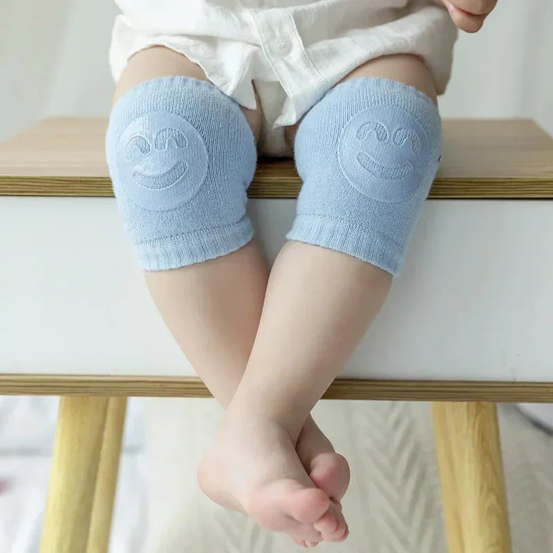 Baby wearing blue knee pads with smiley face design on a wooden stool.